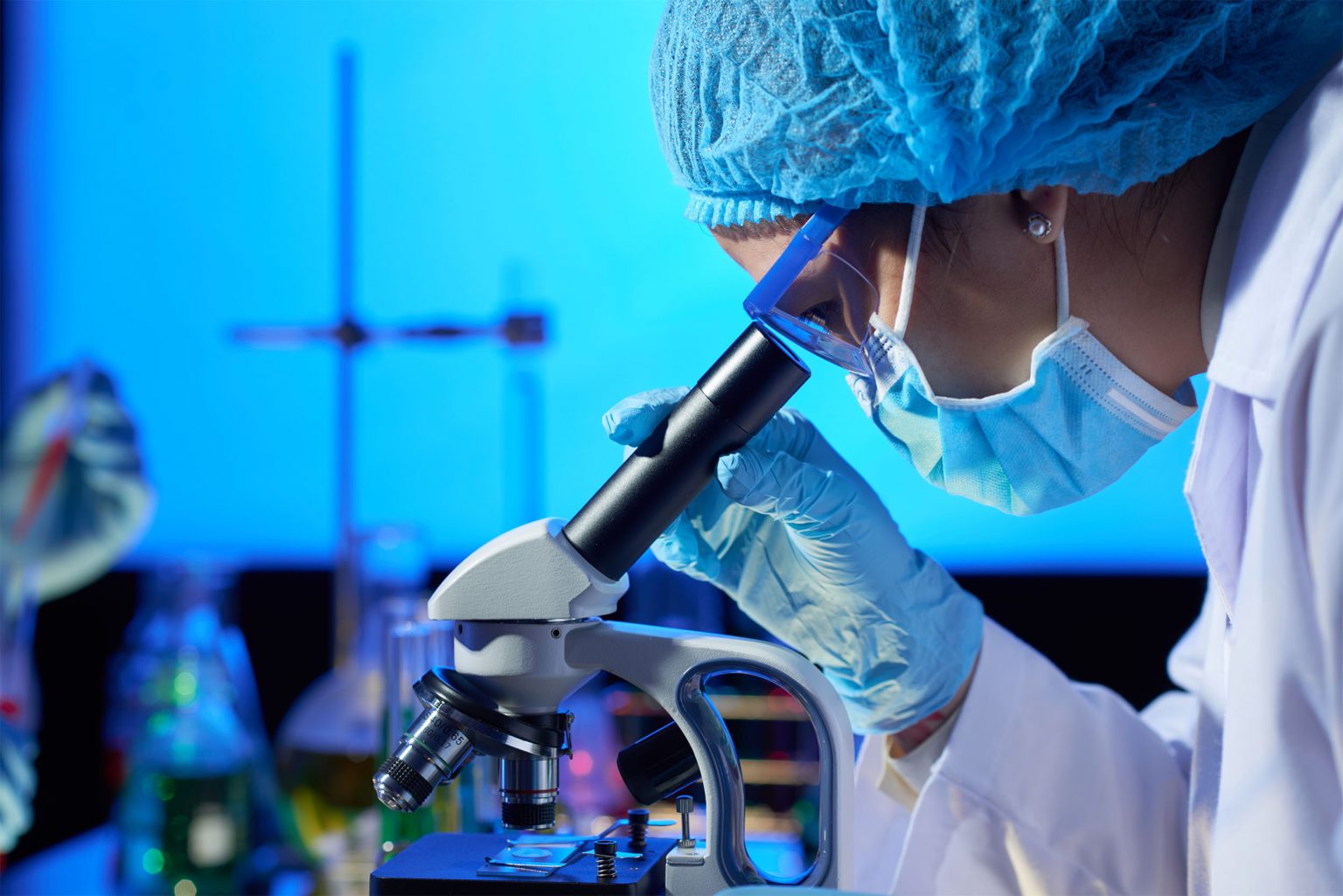 Investigadora con bata blanca y guantes observando una muestra a través de un microscopio de laboratorio